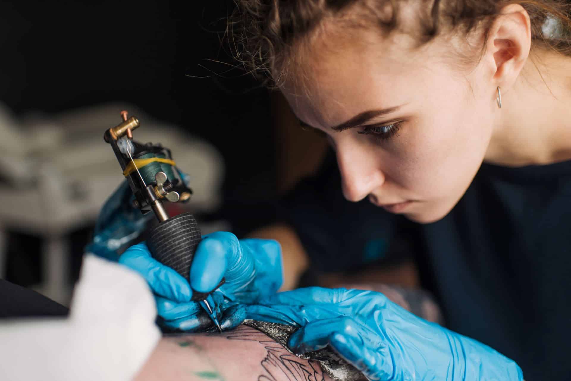 Konzentrierte Tätowiererin mit blauen Handschuhen arbeitet an einem Tattoo und hält die Tattoo-Maschine fest in der Hand.