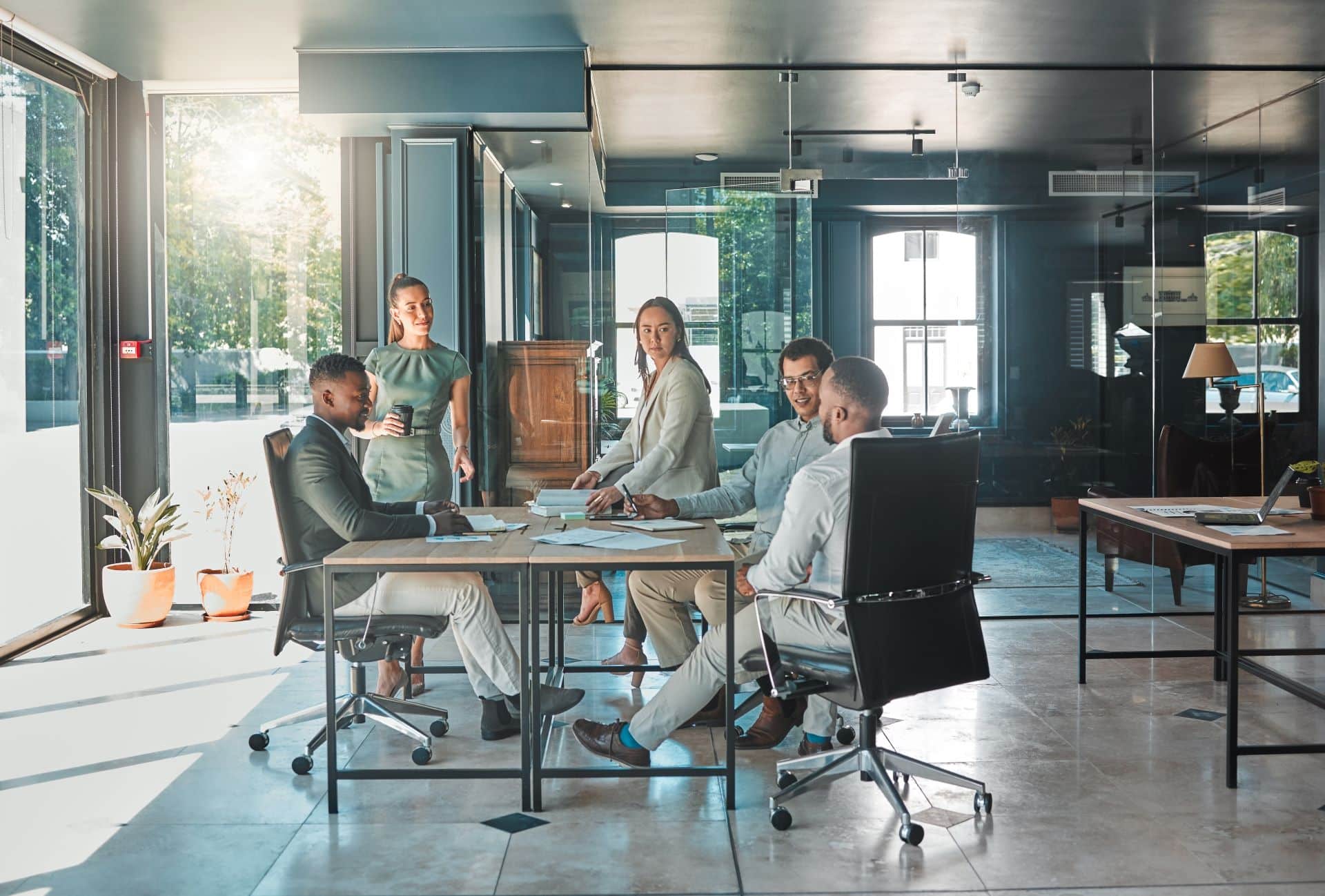 Geschäftstreffen in einem modernen Büro: Eine diverse Gruppe von Kollegen diskutiert um einen Tisch herum, während natürliches Licht durch große Fenster fällt. Die Szene symbolisiert ein professionelles und kooperatives Arbeitsumfeld