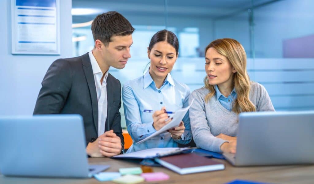 Drei Personen in einer Besprechung analysieren Unterlagen zur Betriebsdatenerfassung und diskutieren Optimierungsmöglichkeiten am Schreibtisch mit Laptops.