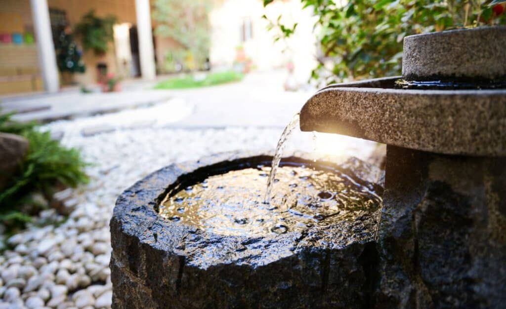 Nahaufnahme eines modernen Wasserelements aus Naturstein über Kies – harmonische Verbindung von Wasser, Licht und Struktur im Garten.